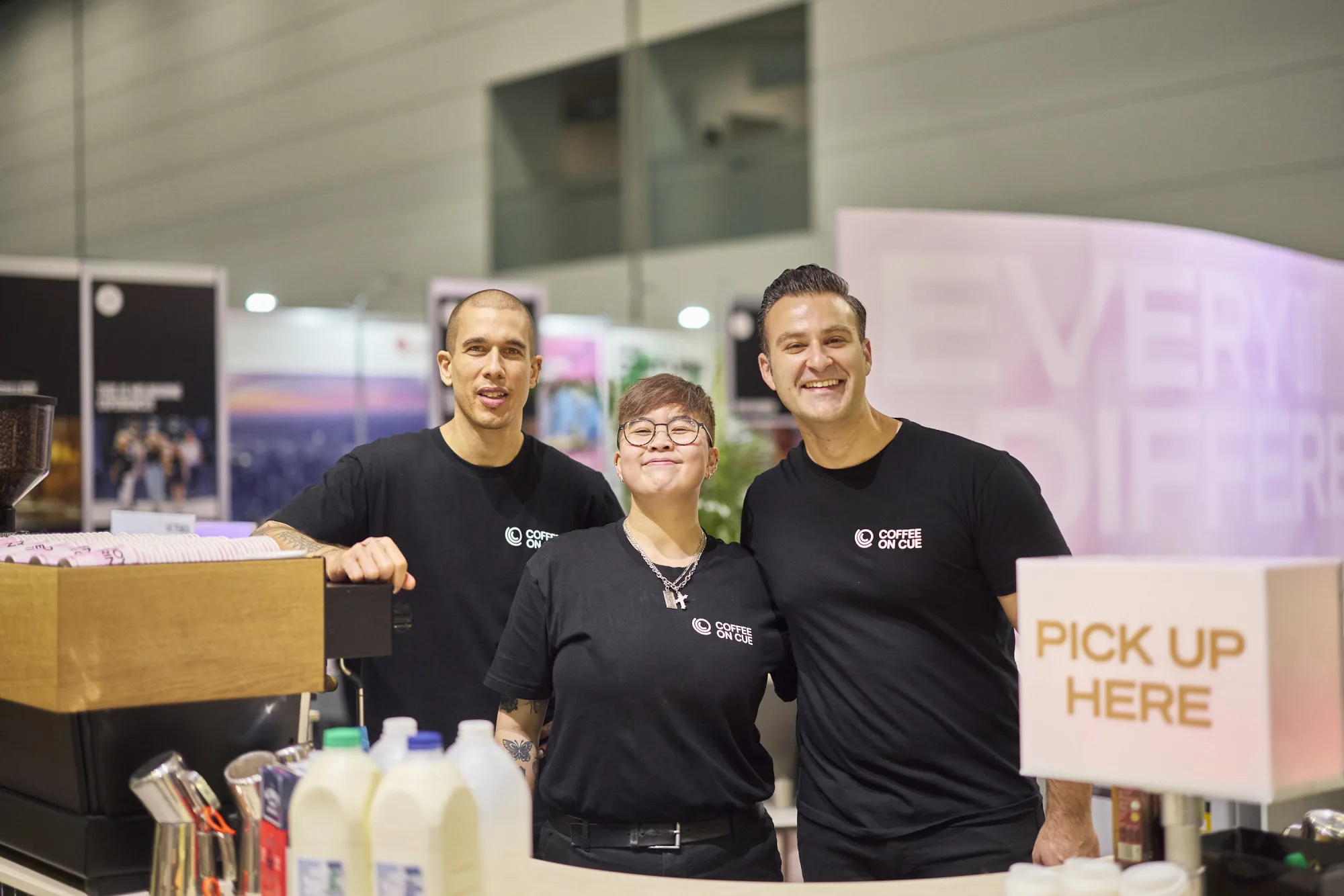 Three Coffee on Cue baristas in black crew shirts smiling behind the espresso machine at a trade-show coffee bar, milk bottles on the counter and a "Pick up here" sign beside them